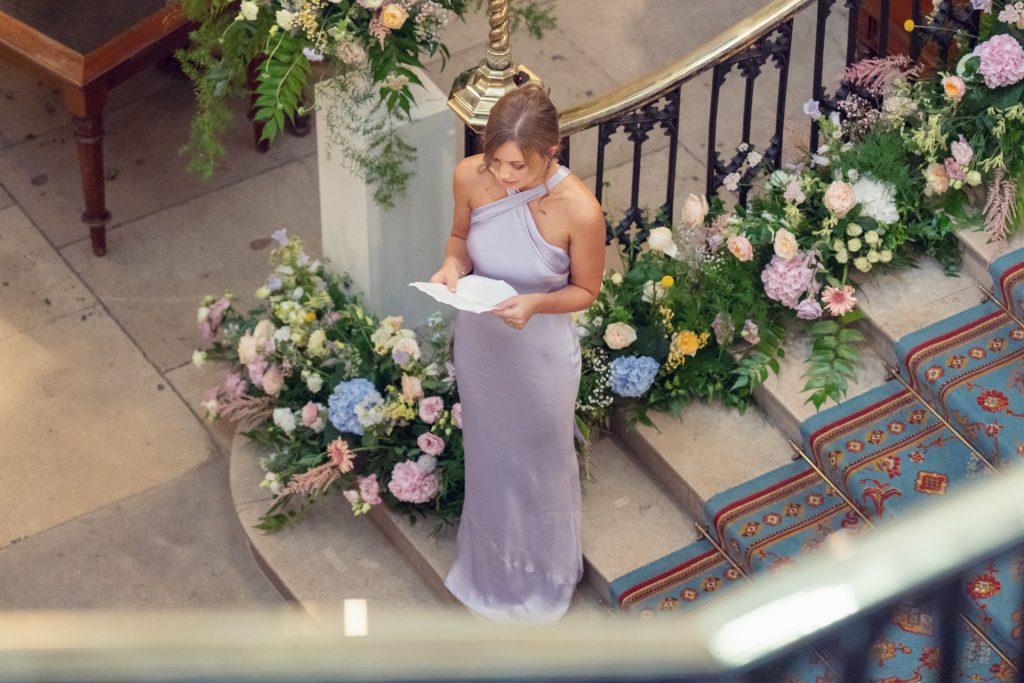 A woman in a long lavender dress stands by a staircase decorated with flowers, holding and reading from a piece of paper at what appears to be a formal event or ceremony.