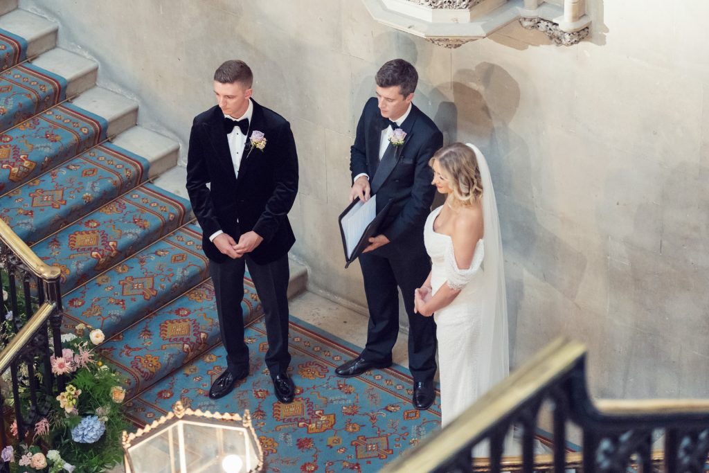 A bride in a white gown and veil stands next to a groom in a black suit and bow tie as a man in a suit holds a folder, officiating their wedding ceremony on ornate carpeted stairs.