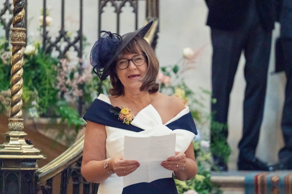 A woman in a formal dress and hat stands indoors, holding papers and smiling. She stands near ornate railings and floral arrangements, suggesting a special event or ceremony.