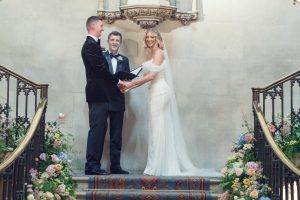 A bride in a white dress and a groom in a black suit stand on ornate steps holding hands, smiling, with an officiant between them. The staircase is decorated with colourful flowers.