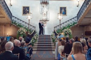 A bride and groom stand at the top of a grand staircase during their wedding ceremony, facing each other and holding hands, with guests seated below in an ornately decorated hall.