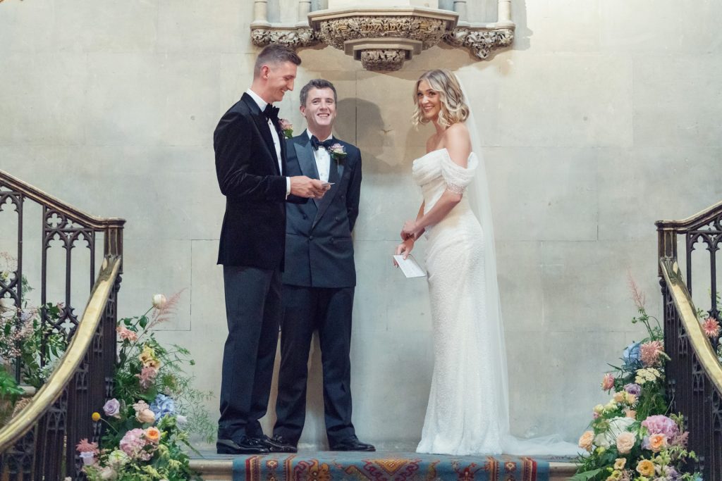 A bride in a white dress and a groom in a black suit stand on a decorated staircase facing each other, holding papers, with an officiant between them and floral arrangements on both sides.