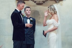 A bride wipes away a tear while facing the groom, who holds a piece of paper. A man in a tuxedo stands behind them, all set against a stone wall, during a wedding ceremony.