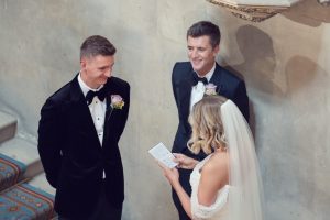 A bride in a white dress and veil reads vows from a note to two men in tuxedos with buttonholes, standing on a staircase with patterned carpet and a stone wall background.
