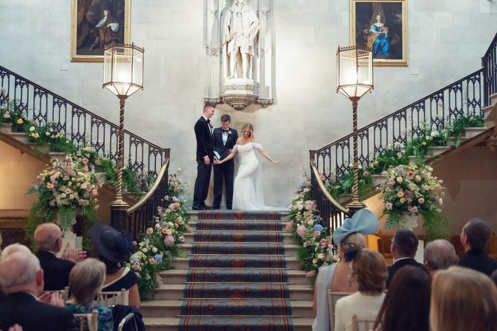 A bride and groom stand holding hands with an officiant at the top of a grand staircase, surrounded by flowers, in front of guests at a formal indoor wedding ceremony.
