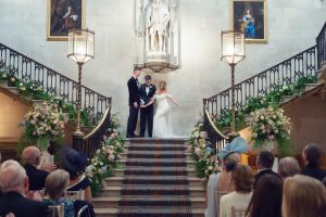 A bride and groom stand holding hands with an officiant at the top of a grand staircase, surrounded by flowers, in front of guests at a formal indoor wedding ceremony.