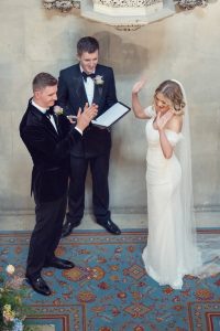 A bride in an off-shoulder wedding dress and veil raises her hand, smiling, while the groom in a black tuxedo applauds. An officiant stands behind them holding a folder, smiling, during a wedding ceremony.