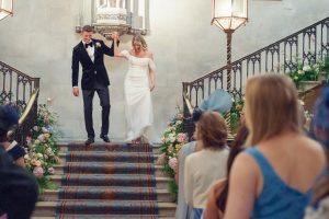 A bride and groom happily walk down ornate stairs decorated with flowers, holding hands, as guests in blue attire watch and smile at their wedding ceremony.
