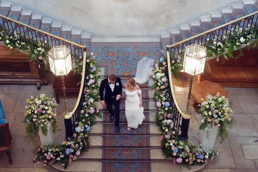 A bride and groom walk down a grand, flower-decorated staircase with ornate railings, holding hands. The bride wears a white dress, and the groom is in a dark suit. The setting is elegant and formal.