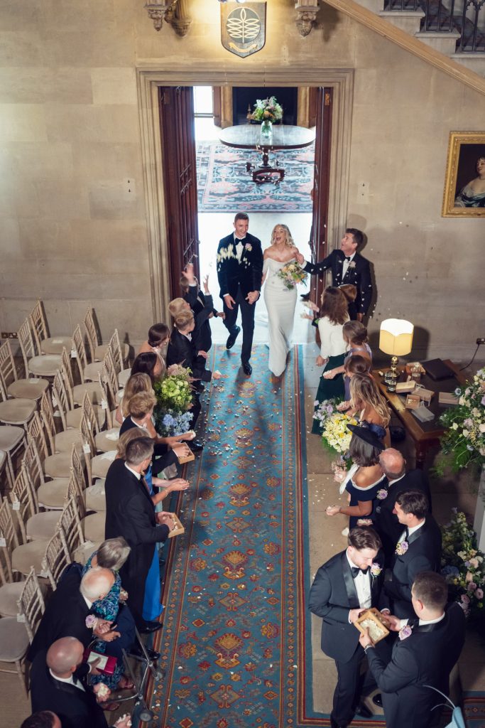 A bride and groom walk down the aisle after their wedding ceremony, smiling and holding hands, as guests on both sides toss flower petals in celebration inside a grand hall with ornate décor.