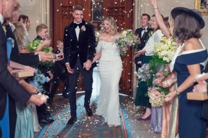 A smiling bride and groom walk hand in hand down an aisle, surrounded by cheering guests who are throwing flower petals. The couple and guests are dressed elegantly and appear to be leaving a wedding ceremony.