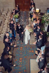 A bride and groom walk down the aisle as wedding guests on both sides throw flower petals. The couple smile, and the guests are seated and standing, dressed in formal attire in a decorated indoor venue.