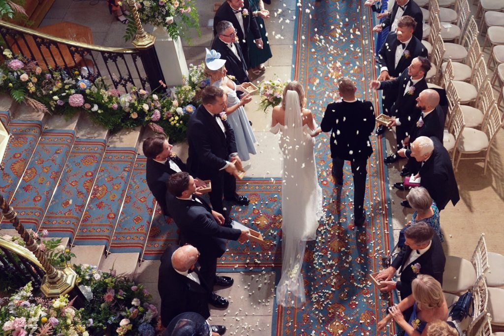 Bride and groom walk down a flower-petal-strewn aisle in a decorated church, surrounded by elegantly dressed guests, some tossing petals in celebration. The scene is viewed from above, showing ornate rugs and floral arrangements.