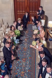Wedding guests dressed in formal attire line both sides of an aisle decorated with flower petals and a blue patterned runner, smiling and applauding as they watch an unseen event at the front of a grand, elegant venue.