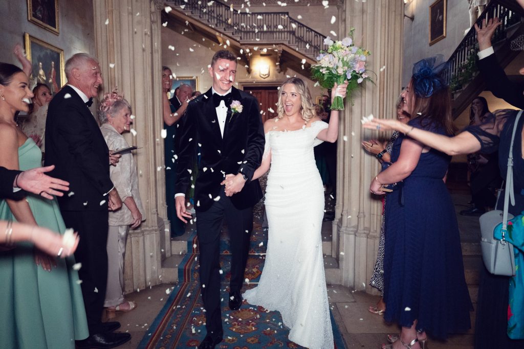 A bride and groom walk down an aisle indoors, smiling and holding hands, as guests throw white confetti and celebrate. The bride holds a bouquet and people on either side clap and cheer in a grand corridor.