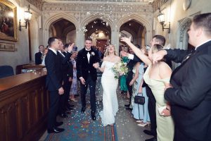 A newly married couple walks down an aisle indoors, smiling and holding hands, whilst guests on both sides cheer and throw flower petals in celebration. The bride wears a white dress and holds a bouquet.