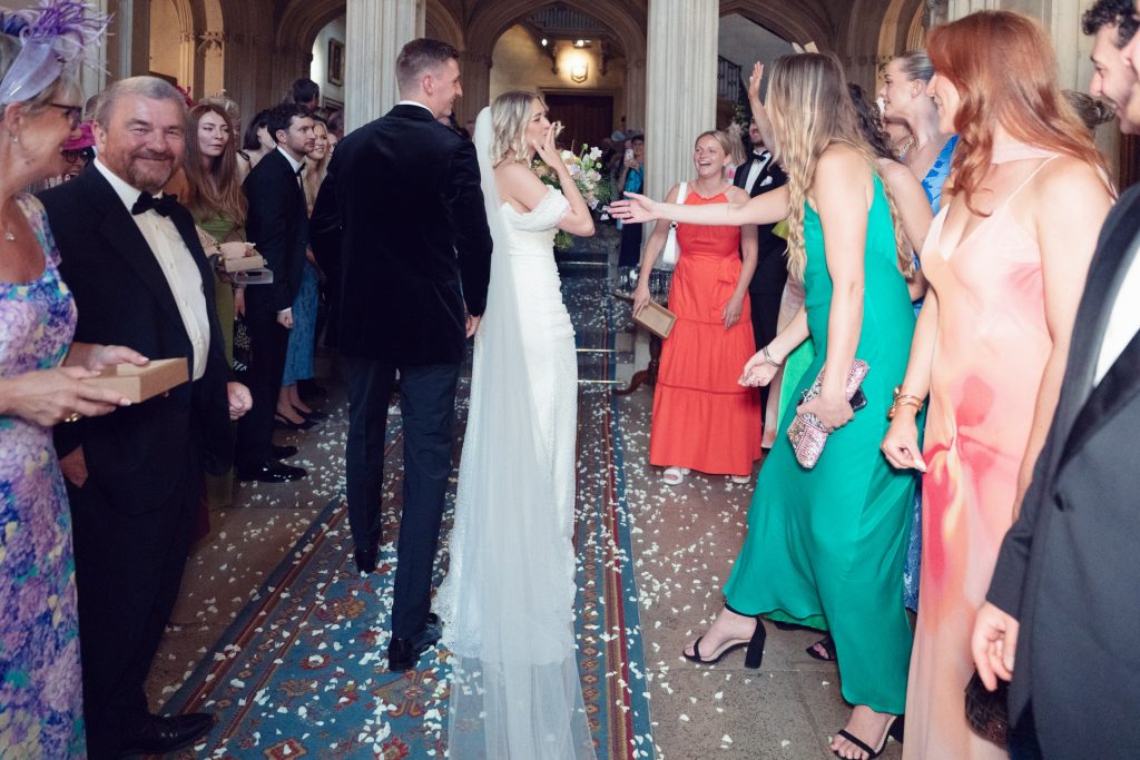 A bride and groom walk down an aisle lined with smiling, well-dressed guests in a grand hall. Flower petals cover the floor as people celebrate and reach out to congratulate the newly-weds.