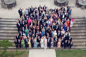 A large group of people dressed formally, including dinner jackets and evening dresses, gathered on outdoor stone steps for a group photo at what appears to be a wedding or formal event. Tables and chairs are visible in the background.