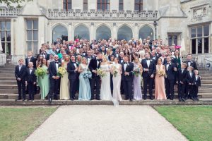 A large wedding party poses on stone steps in front of a grand historic building. The group includes people in formal attire, bridesmaids in pastel dresses, groomsmen in black suits, and flower girls and boys.
