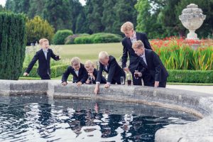 Six boys dressed in suits gather around a stone fountain, with some leaning over to touch the water. Lush gardens and greenery surround them, and a few boys hold glasses with dark beverages.