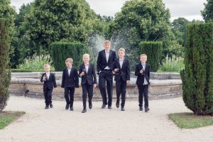 Five boys in suits walk together in front of a garden fountain, smiling and holding fizzy drink cans. Lush green trees and neatly trimmed bushes surround them on a bright, cloudy day.