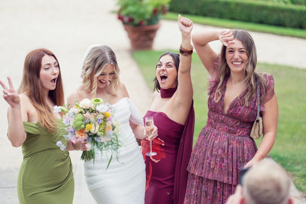 Four women pose happily at an outdoor event, with one in a white wedding dress holding a bouquet. The others wear colourful dresses and make celebratory gestures, smiling and laughing together. Greenery is visible in the background.