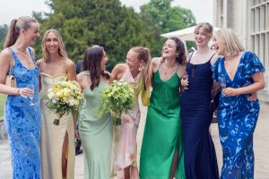 Seven women dressed in colourful, elegant dresses stand outdoors, smiling and laughing together. Two hold bouquets of flowers, and another holds a glass, suggesting a celebratory or formal event. Trees and a building are visible in the background.