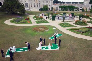 A bride and groom play crazy golf with guests on a lawn outside a large, historic building surrounded by formal gardens and flowerbeds, whilst more people gather near a circular fountain.