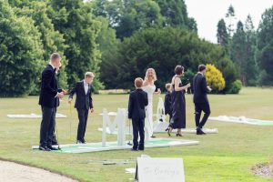 A group of people dressed formally play mini-golf on a lawn, surrounded by greenery. A white wedding sign with The Berkleys is visible in the foreground. The mood is light and celebratory.