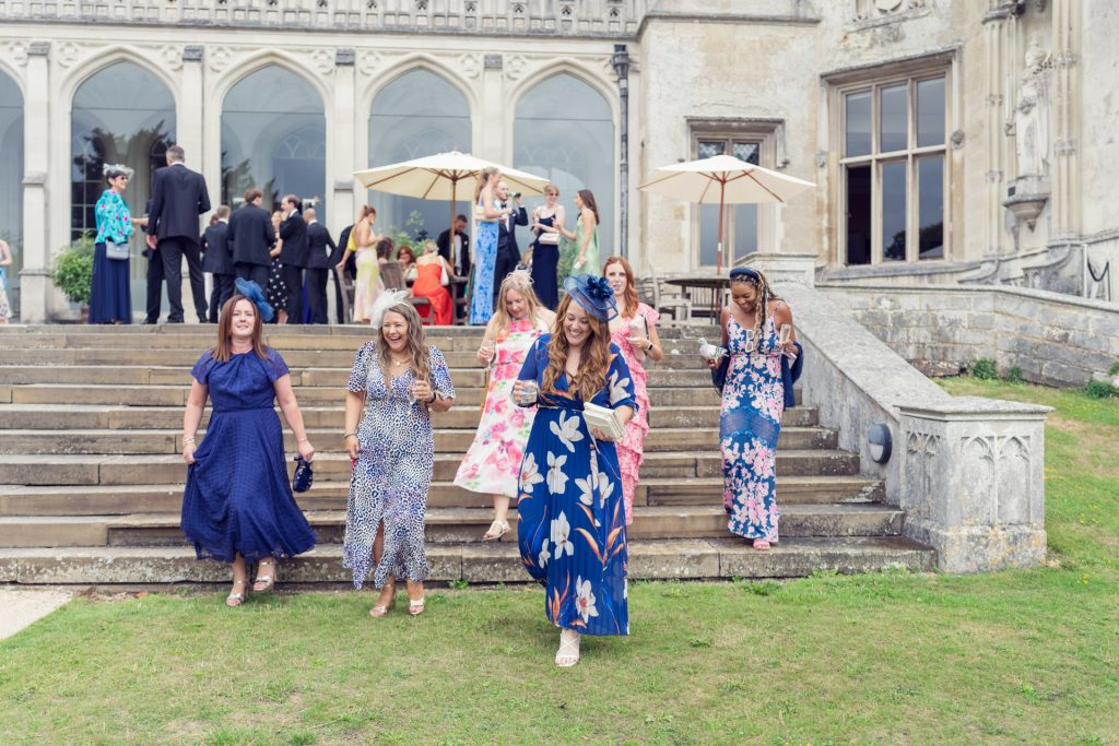 A group of women in colourful dresses and hats walk down stone steps outside a grand historic building during a formal outdoor event, with other guests mingling in the background.