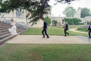 A bride in a white dress runs up stone steps outside, followed by three men in black suits across a grassy path, near a historic stone building and gardens.