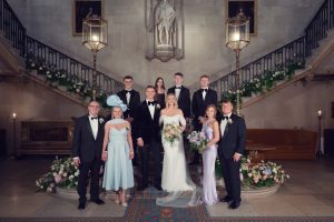 A wedding party poses indoors on a grand staircase. The bride, in a white dress holding a bouquet, stands beside the groom in a black tuxedo, surrounded by bridesmaids, groomsmen, and family members in formal attire.