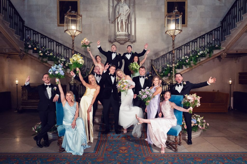 A wedding party poses joyfully in a grand hall with twin staircases, floral decorations, and elegant attire. Bridesmaids wear pastel dresses and groomsmen wear tuxedos, all smiling and waving for the camera.