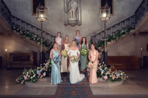 A bride in a white gown stands with seven bridesmaids in pastel dresses on a grand staircase decorated with flowers inside an elegant hall. Each woman holds a bouquet and smiles at the camera.