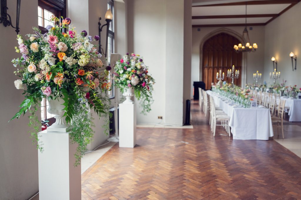 A bright, elegant banqueting hall with tall floral arrangements on pedestals and long tables laid for a formal event. The room features large windows, wooden floors, and candle-lit chandeliers.