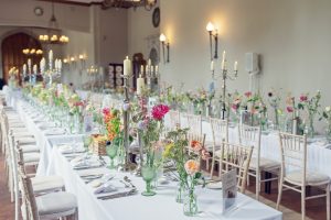 Long banquet tables with white tablecloths are set for an event, decorated with colourful flowers in glass vases, tall candelabra, and neatly arranged chairs in an elegant, well-lit hall.
