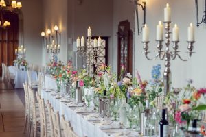 A long, elegant banquet table is set with white linen, tall silver candelabra with lit candles, glassware, plates, and colourful floral arrangements, creating a festive atmosphere in a softly lit room.