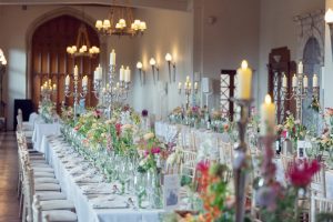 A long banquet table set for an event, decorated with tall candelabra, lit candles, glassware, and colourful flower arrangements in a grand, elegant hall with chandeliers and arched wooden doors.