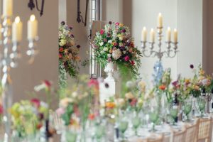 A long dining table elegantly set with glassware and colourful flowers, with tall floral arrangements and lit candelabra in a bright, airy room.