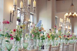 Elegant banquet table set with tall silver candelabra holding lit candles and glass vases filled with colourful fresh flowers; green glassware and white chairs complete the sophisticated setting.