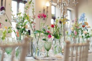 A close-up of a banquet table elegantly set with glassware and green goblets, decorated with tall, colourful wildflowers and candles in a bright, softly lit room.