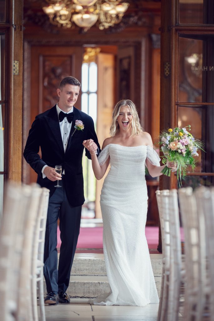 A smiling bride in a white dress and a groom in a black tuxedo with a drink walk down an aisle indoors, with the bride holding a bouquet and looking joyful. Elegant decor and chairs line the aisle.
