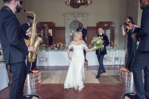 A bride and groom joyfully enter their wedding reception, holding hands. Musicians play saxophone, guitar, and sing on raised platforms around them whilst guests watch from banquet tables in a grand hall.