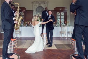 A bride in a white dress dances with a groom in a black suit. Musicians play saxophone and trumpet on podiums nearby. The scene is set in an elegant hall with flowers and a long table in the background.