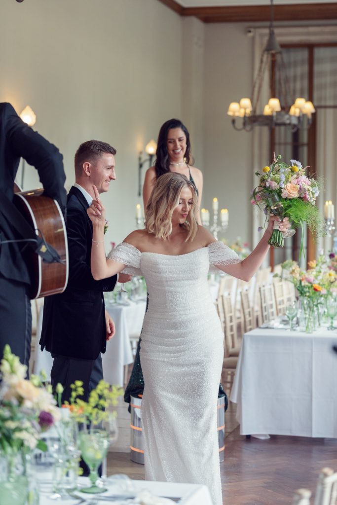 A bride in a white dress holds a bouquet and raises her arm whilst walking beside a smiling groom in a suit at a decorated wedding reception. Guests and floral arrangements are visible in the background.