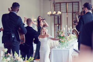 A bride in a white dress joyfully dances at a wedding reception with live musicians playing and a woman singing near a long, flower-decorated table in a bright, elegant room.