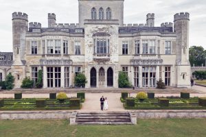 A bride and groom stand holding hands in front of a large, historic stone manor with manicured gardens and symmetrical landscaping.
