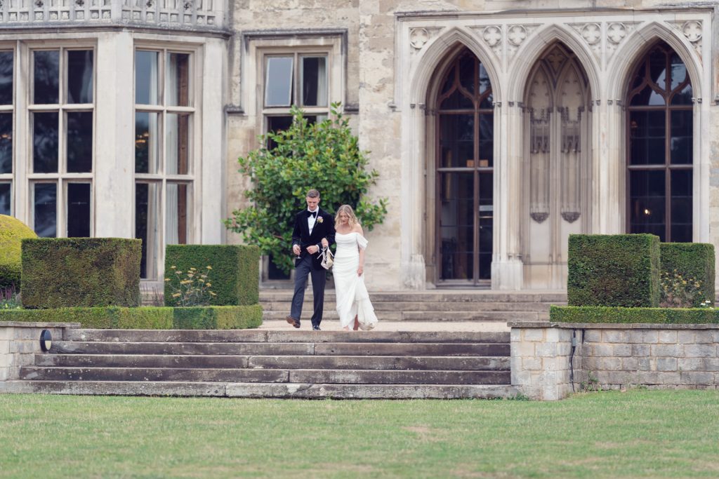 A bride and groom walk down stone steps outside a grand historic building with large windows and ornate arches, surrounded by manicured hedges and a lawn.