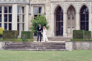 A bride and groom walk down stone steps outside a grand historic building with large windows and ornate arches, surrounded by manicured hedges and a lawn.
