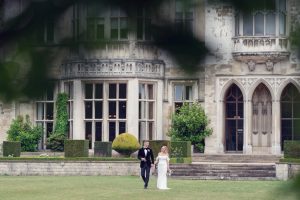 A bride and groom walk hand in hand on a lawn in front of a grand, historic stone building with large windows and arched entrances, framed by tree branches in the foreground.